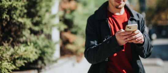 Fototapeta premium A young male outdoors interacting with his smartphone, casually dressed in a red shirt and black jacket with a cheerful expression.