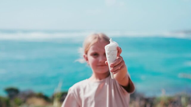 Close-up of a young girl smiles proudly while showing off a seashell she found on the sandy beach, with ocean waves in the background.