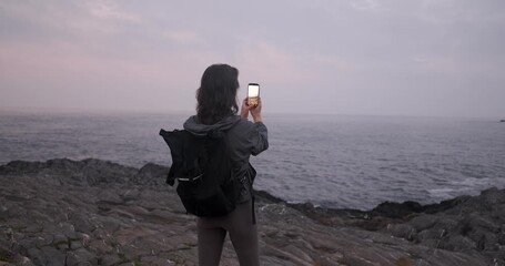 Young adult woman with backpack takes photos of the rough sea with her phone while standing on rocky cliffs at dusk or dawn.