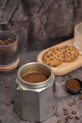 Geyser coffee maker with ground coffee standing on the table. Coffee beans in a glass. Coffee in a black spoon. Oatmeal cookies with chocolate chips on a wooden board. Dark background. Still life