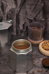 Geyser coffee maker with ground coffee standing on the table. Coffee beans in a glass. Coffee in a black spoon. Oatmeal cookies with chocolate chips on a wooden board. Dark background. Still life