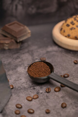 Ground coffee in a black spoon. Pieces of broken dark chocolate. Spilled coffee beans. Oatmeal cookies on a wooden board. Dark background. Still life