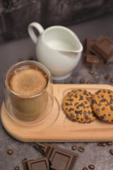 Close up. Coffee in a glass and oatmeal cookies on a wooden board. Milk jug on the table. Pieces of broken dark chocolate. Spilled coffee beans. Dark background. Coffee service