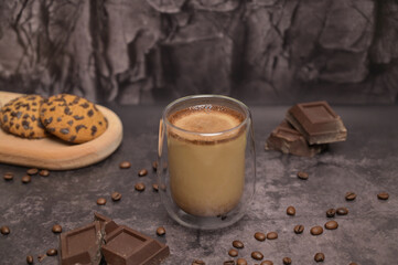 Coffee in a transparent glass. Oatmeal cookies on a wooden board on the table. Pieces of broken dark chocolate. Spilled coffee beans. Dark background. Coffee service. Still life