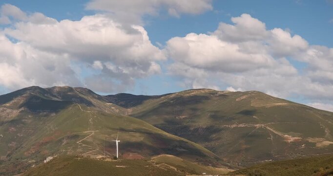 Slow motion shot of a green mountain range with a single wind turbine under a blue sky with fluffy white clouds moving slowly.