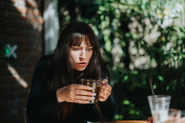 A woman wearing a black sweater appears surprised while holding a drink, sitting outdoors in a...
