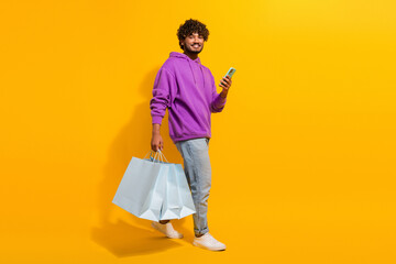 Cheerful young man in a purple hoodie holding shopping bags and a smartphone against a vibrant...