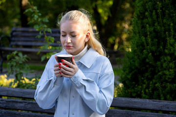 Young blonde woman with red nails holds a black paper cup close to her face, eyes closed, savoring the warmth and aroma of a hot drink on a sunny morning in a green city park