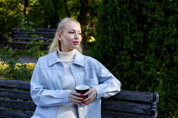 Stylish young woman with light blonde hair and red nails holds a black paper coffee cup while sitting on a wooden bench in a quiet park, surrounded by lush greenery on a cool autumn day