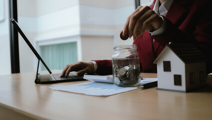 Businesswoman analyzing financial charts with a jar of coins and a small house model, representing investment, savings, and financial planning.