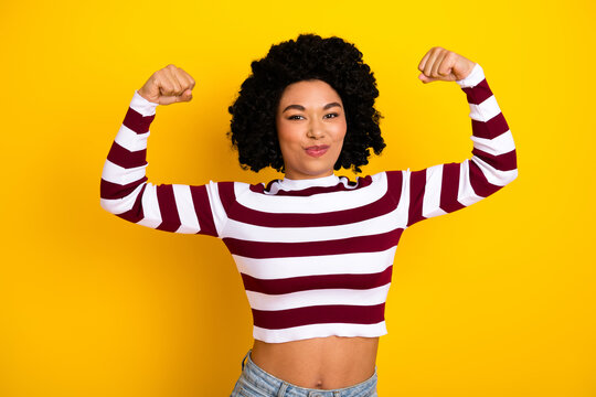 Fototapeta Confident woman flexing arms in striped top against vibrant yellow backdrop showcasing style and strength