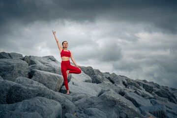 Woman wearing red sportswear doing a yoga tree pose on rugged rocks