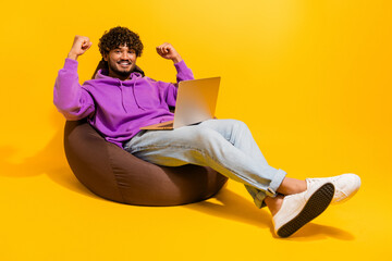 Happy young man with curly hair sitting on a bean bag and celebrating success while using a laptop...