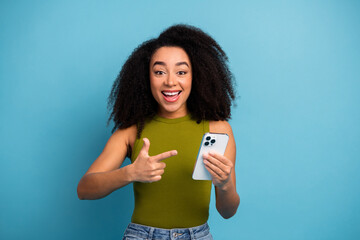 Excited young woman holding a smartphone and pointing, cheerful expression against a vibrant blue...