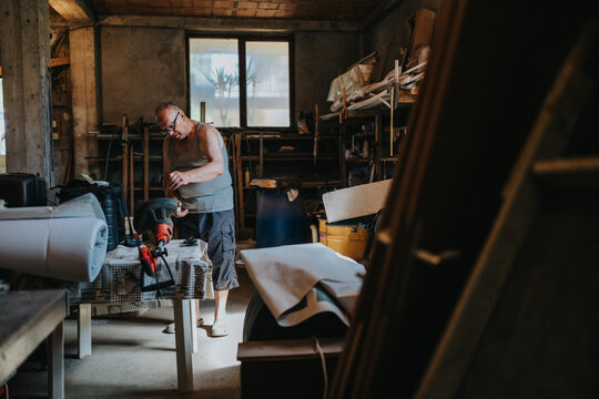 A middle-aged man in a tank top operates a grinder in a messy workshop filled with tools and materials. Bright light from a window highlights the focused, hands-on craft activity. - Powered by Adobe
