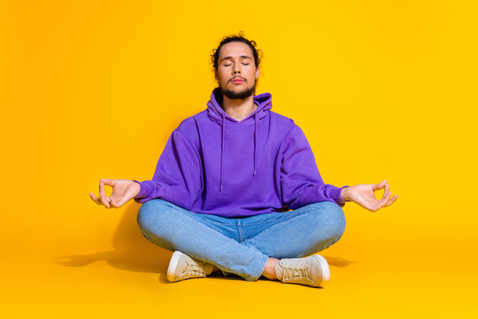 Young man in a peaceful meditation pose wearing a purple hoodie, seated cross-legged against a vivid yellow background