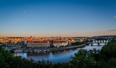 Panorama landscape with evening sunlight bathes Prague Old Town, Czech Republic, with the Vltava River below and the moon rising in a clear sky.