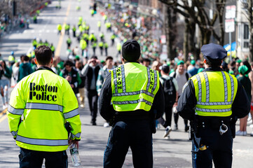 Three Boston police officers in neon safety vests watching the St. Patrick’s Day parade down the street. Boston, MA, USA - March 17th, 2024