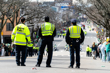 Three Boston police officers in neon safety vests watching the St. Patrick’s Day parade down the street. Boston, MA, USA - March 17th, 2024 © Konrad