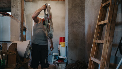A muscular man in a gray tank top works in a cluttered workshop, lifting a coil of wire while surrounded by ladders and various hardware, capturing hands-on labor and preparation.