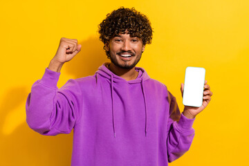 Happy young man in vibrant hoodie celebrates success while showcasing a smartphone on a bright...