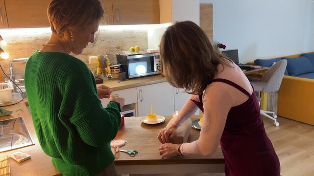 Friends preparing present at kitchen island under Happy Birthday garland and string lights, cozy indoor celebration setup.