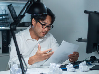 Focused Asian Businessman looking at business report paper document with confusion and frustration, dealing with difficult task and decision on financial problem sit at office desk in workplace night.