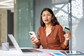 Woman holding a credit card and telephone, looking shocked and experiencing a payment error or fraud issue while trying to make an online purchase using a laptop