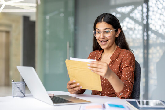 Young woman sitting at a desk in an office setting, joyfully opening an important envelope, reacting with a surprised and happy expression to positive news or results
