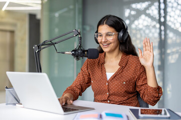 Smiling indian woman wearing headphones and glasses sitting at a desk, using a laptop and a professional microphone to record a podcast or conduct a video call