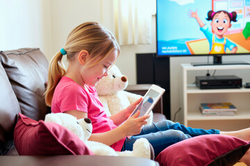 Caucasian girl child sitting on sofa using digital tablet surrounded by stuffed animals, watching animated educational program on digital tablet in living room, engaging with technology