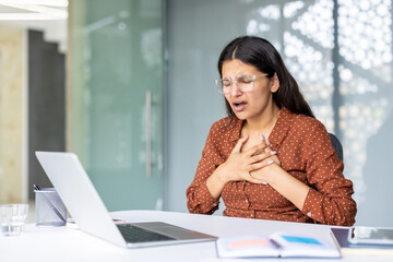Young indian woman experiencing sudden severe chest pain or heart problems while sitting at her office desk, holding her chest with both hands and expressing discomfort and suffering
