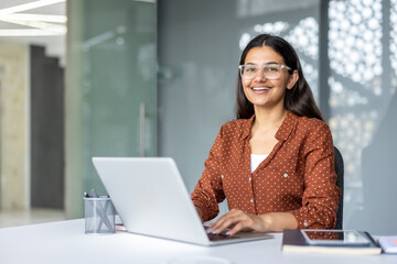 Indian businesswoman smiling confidently. Wearing glasses. And typing on a laptop while sitting at a clean desk in a modern corporate office environment. Representing professionalism and productivity