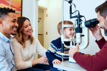 Caucasian boy sitting at ophthalmology device receiving eye exam from male optometrist while smiling parents, young Caucasian woman and young Black man, watching and supporting nearby