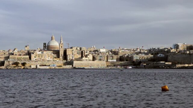 Valletta skyline, Malta. A panoramic view of the historic fortifications from a stone pier on the waterfront. A blend of ancient history and deep blue Mediterranean water under a dramatic sky.