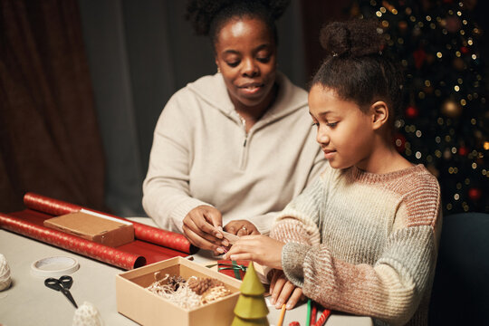 Black woman helping Black girl wrapping Christmas gifts at table, both focused on decorating present with festive materials, Christmas tree with ornaments visible in background