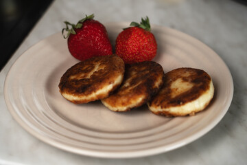 On a plate, pancakes filled with cottage cheese are served with fresh strawberries on the side. The pancakes are being cooked in a frying pan.