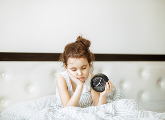 Cute little kid sleepy girl sitting on bed waking up in morning and holding alarm clock.