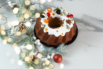 Christmas food still life with iced fruit cake, holly, snow covered winter greenery on light background.