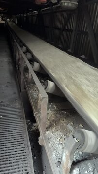 A dynamic, close-up shot captures a heavy industrial conveyor belt transporting a high volume of crushed material, such as cement or limestone, at great speed through a dark factory interior. The stee