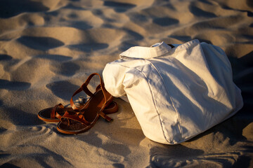Minimalist summer vacation concept. White canvas tote bag and brown leather sandals resting on soft sand dunes during golden hour sunset light.
