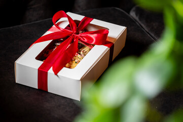 Gift box with assorted decorated treats, tied with red satin ribbon, placed on dark fabric with blurred green plant in foreground. Ideal for celebration, holiday, or product presentation themes.