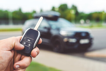 Close-up of a man unlocking a car using a key fob, with a black car parked in a car park in the background.