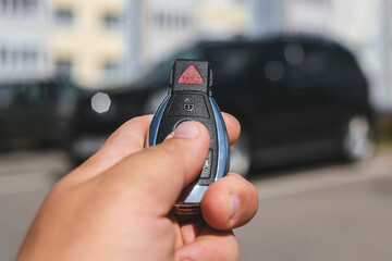Close-up of a man unlocking a car using a key fob, with a black car parked in a car park in the background.
