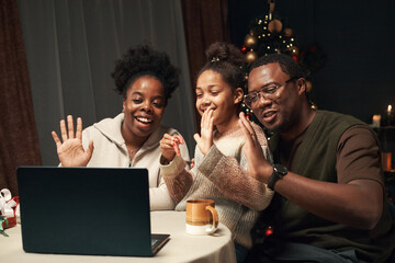 Black woman, Black man, and Black child sitting together waving at laptop screen during Christmas, smiling and engaging in video call with decorated Christmas tree in background