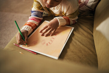 Black child drawing candy cane on paper using colored pencils while lying on couch, hands and face partially visible, celebrating Christmas with creative holiday activity