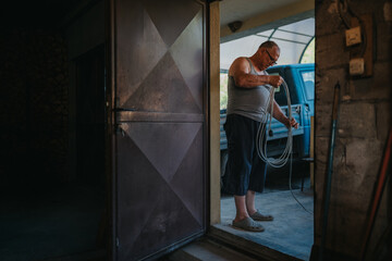 A man in a gray sleeveless shirt and shorts winds a hose outside a garage, with a blue van visible...