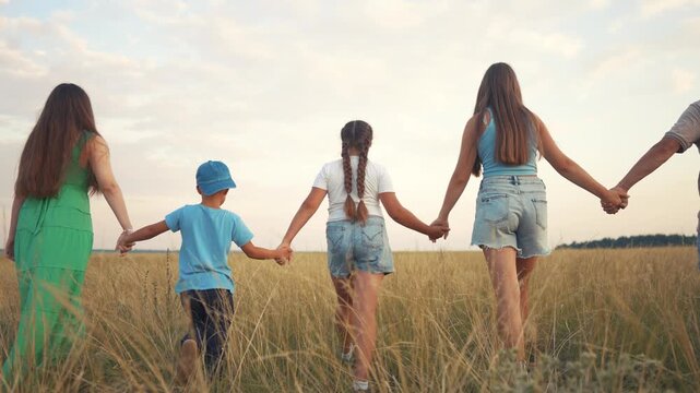 A woman and girl hold hands, walking through the field as the man and boy follow behind. The girl leads the boy and woman deeper into the grassy field under the sky.