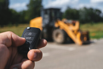 A close-up of a man opening a construction vehicle with a key fob, with a yellow forklift parked in...