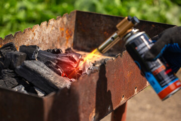 Close-up of a hand in a glove, using a butane torch to quickly start fire and ignite black charcoal briquettes in a rusty metal BBQ grill